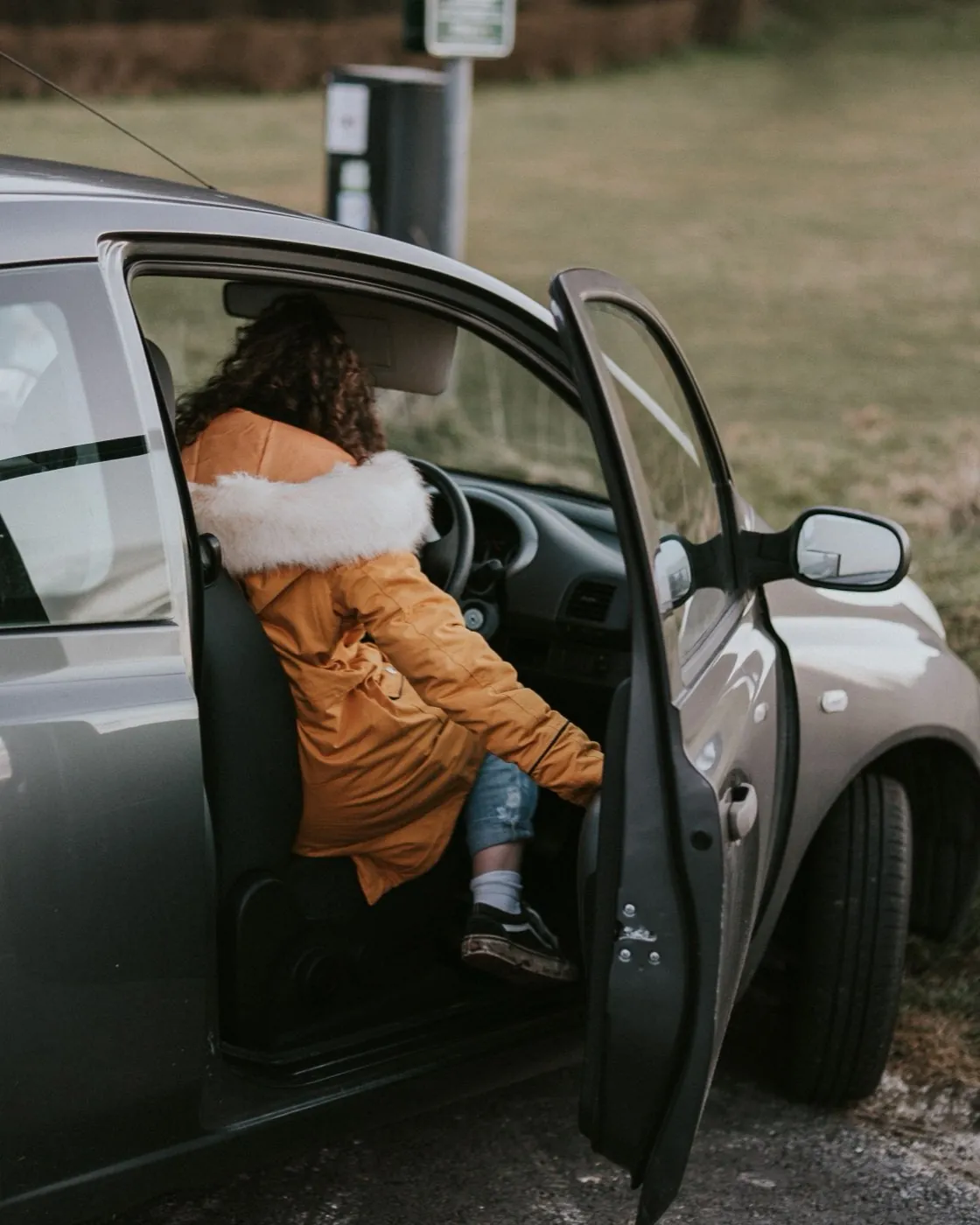 A woman in a mustard parka getting into the driver's seat of her silver hatchback