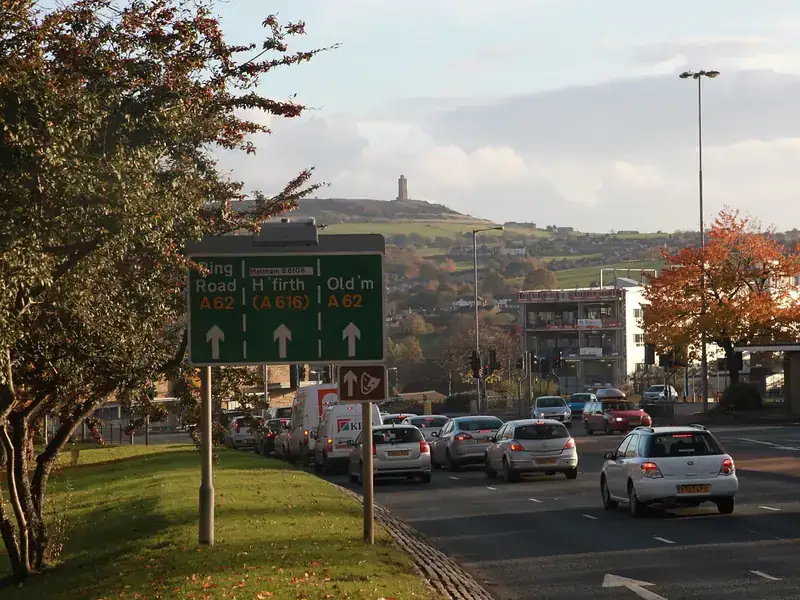 Traffic on the Huddersfield ring road below a sign for the A62 and A616, with Castle Hill's Victoria Tower on the skyline