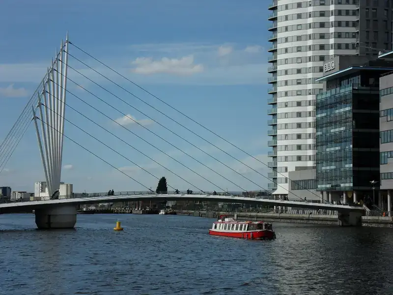 The cable-stayed footbridge at MediaCityUK on Salford Quays, with a red boat on the water and tall buildings beside it