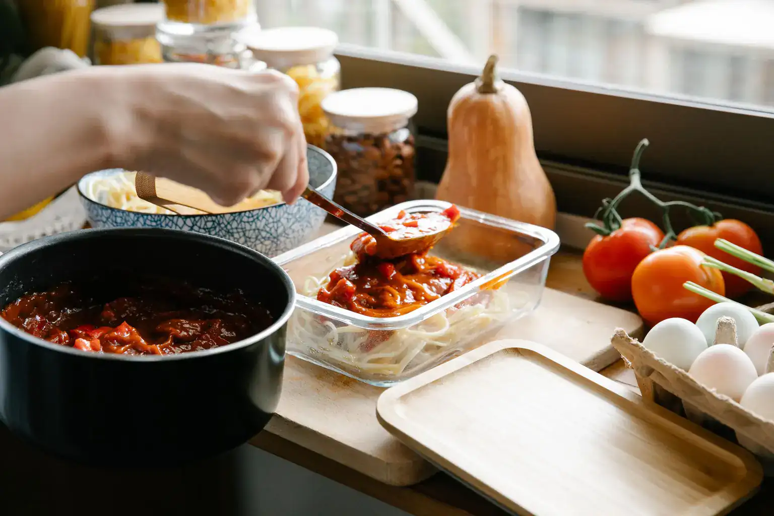 Homemade tomato pasta sauce being spooned into a glass container of spaghetti on a kitchen worktop