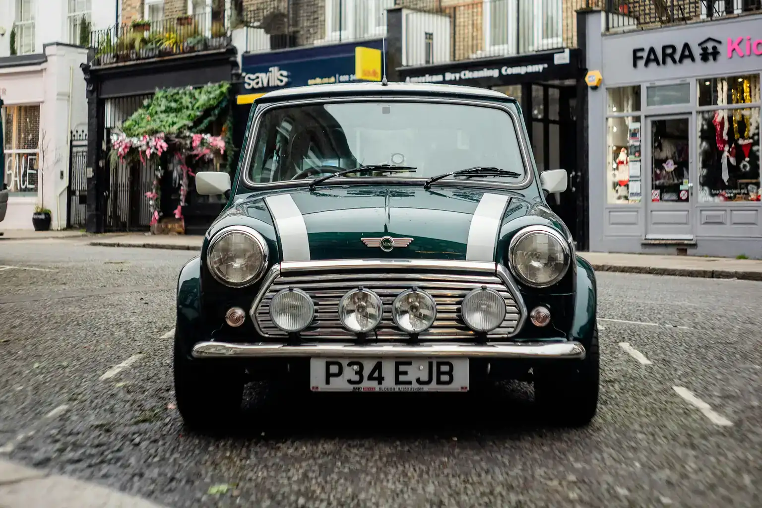 A classic dark green Mini with white bonnet stripes parked on a British high street