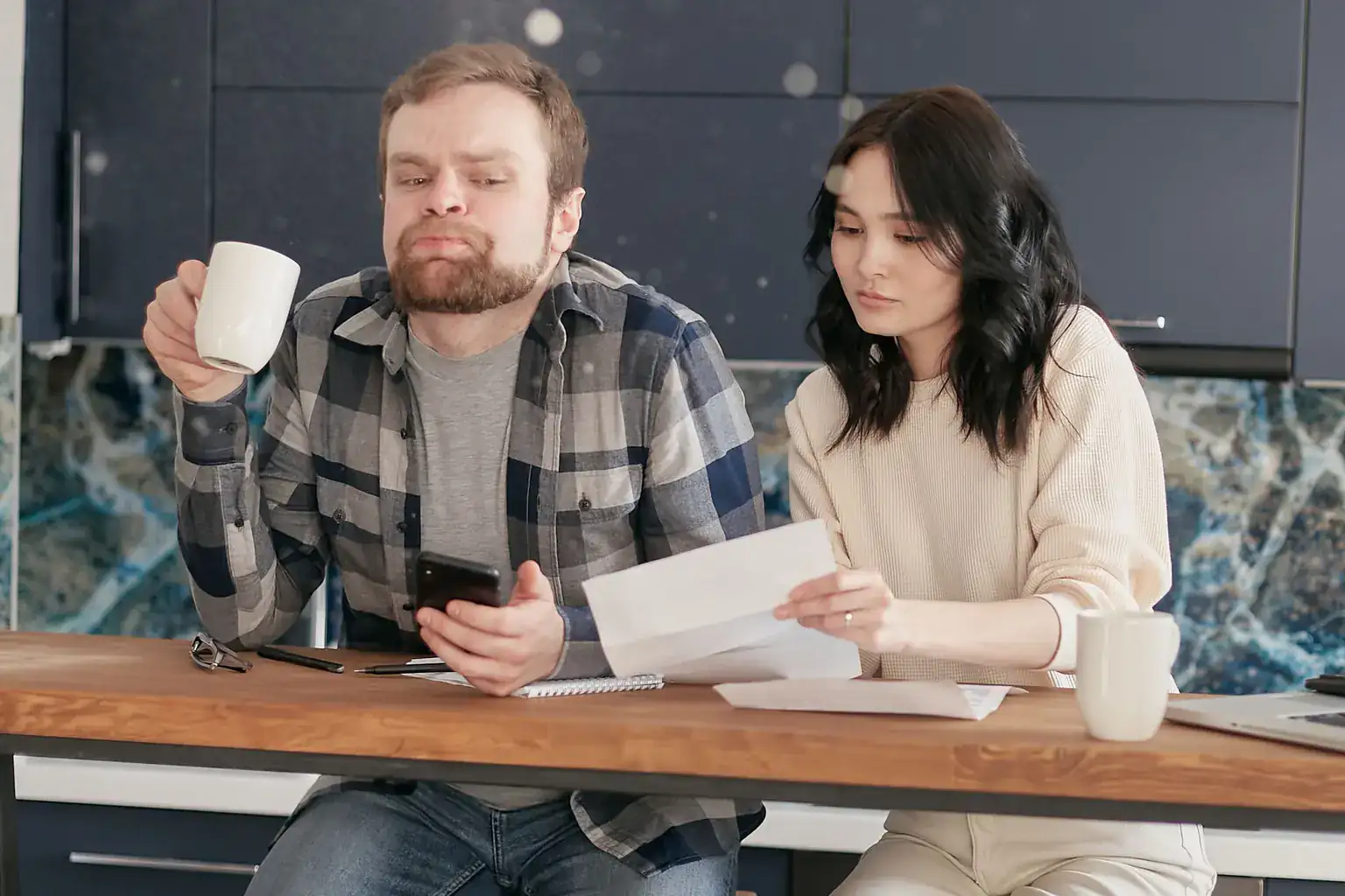 A couple at a kitchen table go through bills and letters together, one checking a phone