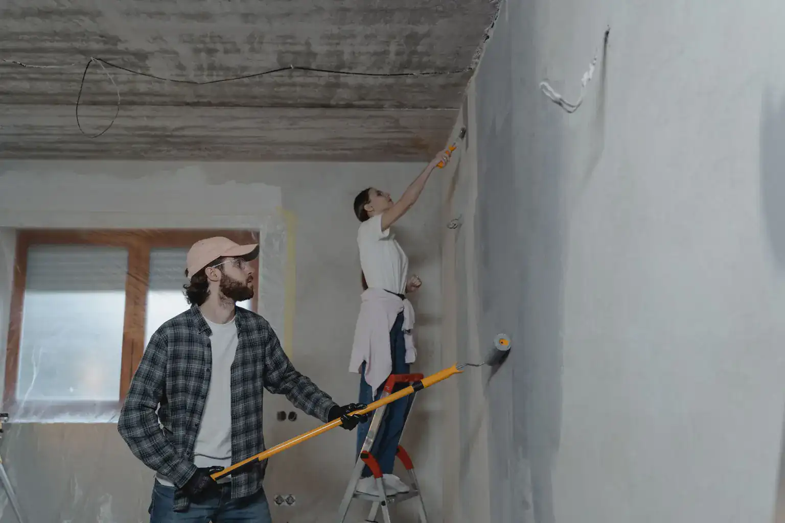 A couple painting freshly plastered walls with a roller and brush during a home renovation