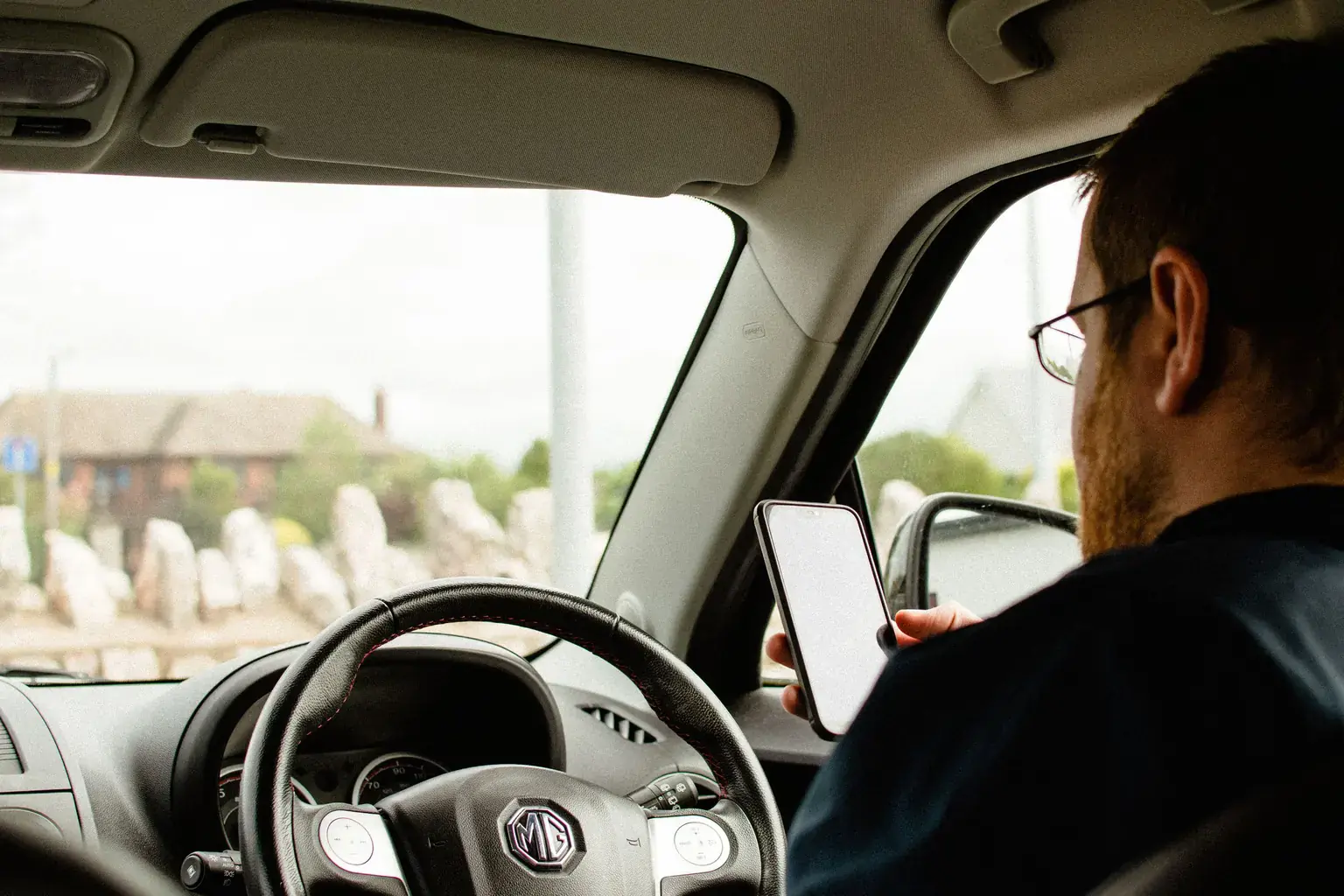 A man in glasses sits behind the wheel of his parked right-hand-drive car checking his phone