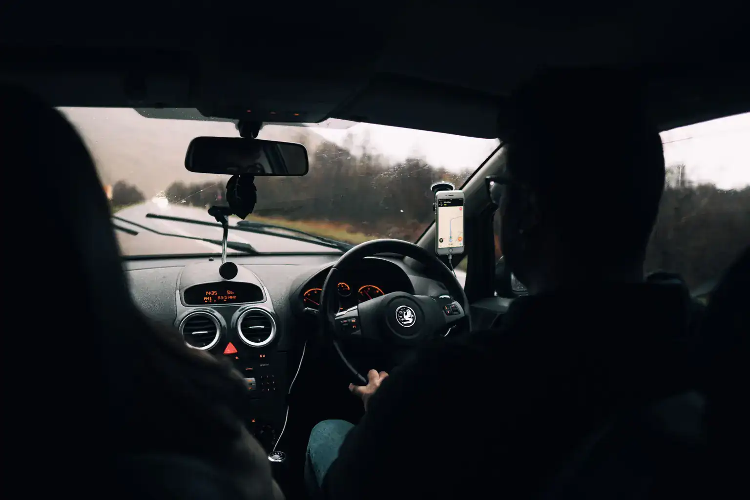 A man driving his right-hand-drive hatchback along a wet country road, seen from the back seat