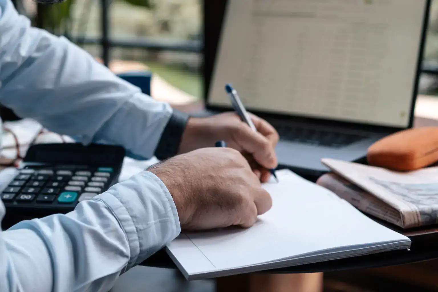 A man writes figures in a notebook beside a calculator and a laptop showing a spreadsheet