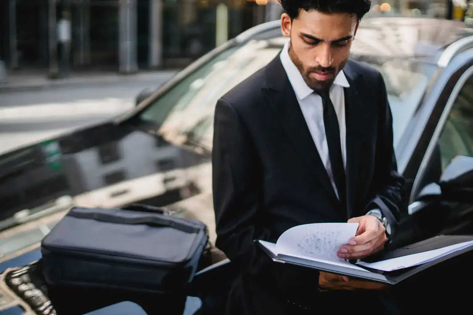 A man in a suit reads through paperwork while leaning against his car