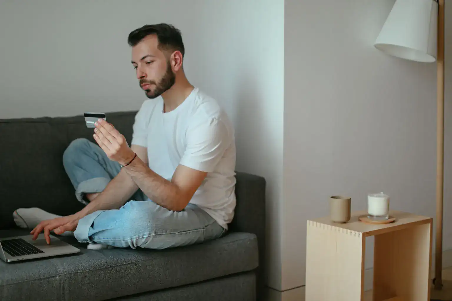 A man on a sofa holds up a credit card while buying something on his laptop