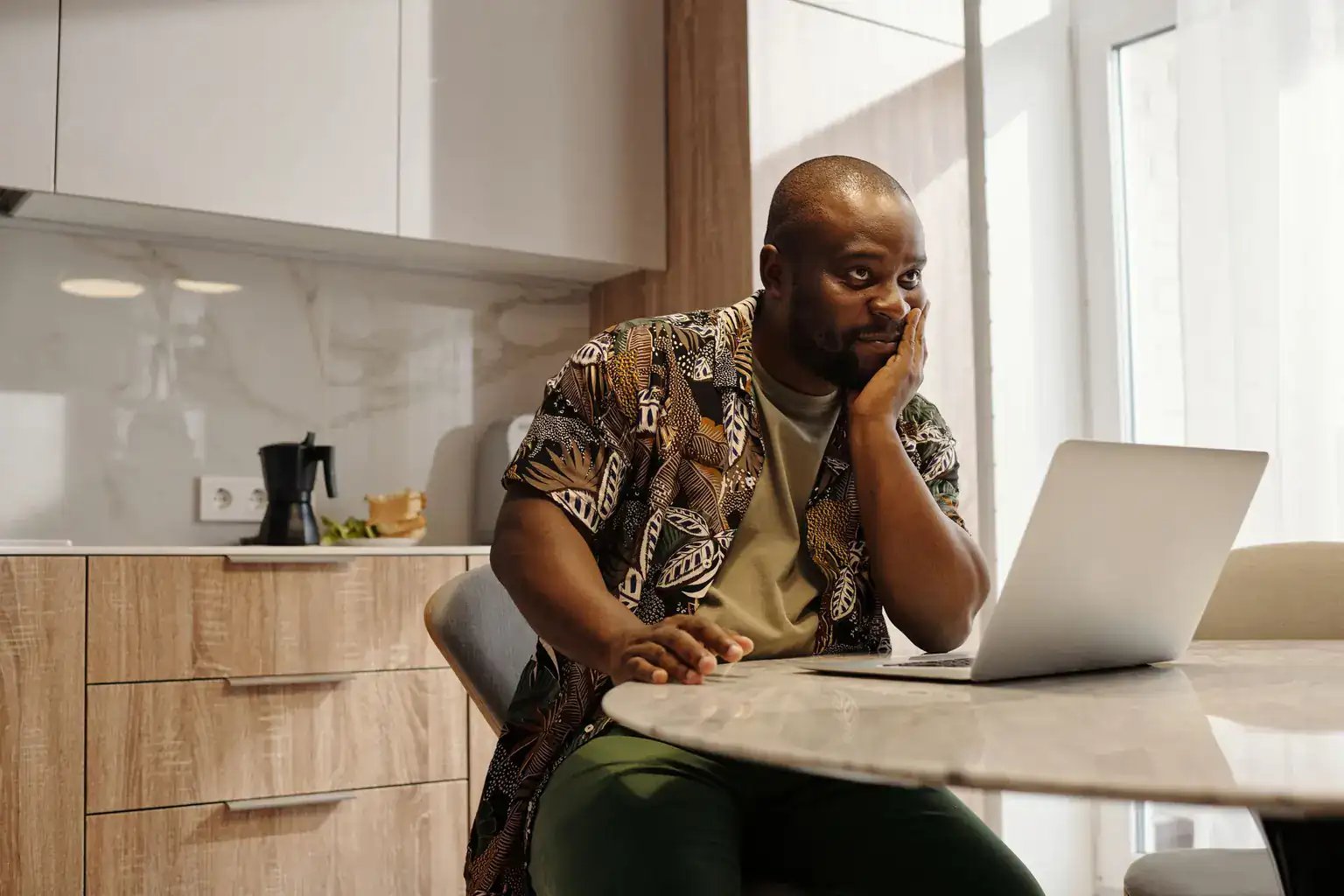 A thoughtful man rests his chin on his hand, looking at a laptop in his kitchen