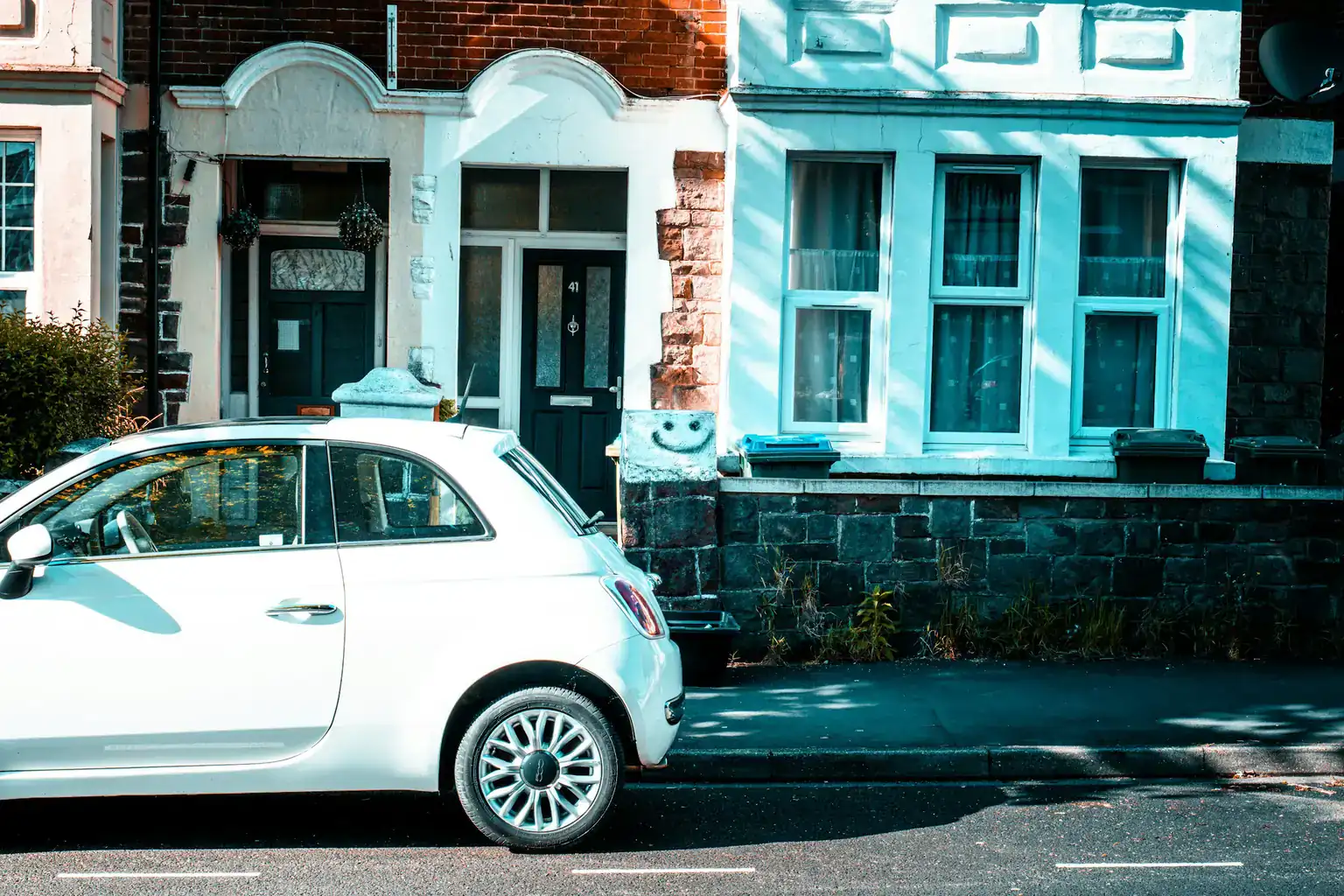 A small white hatchback parked at the kerb outside a pair of British terraced houses