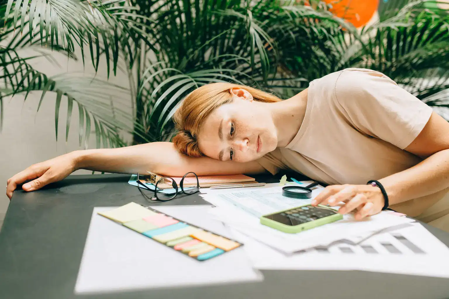A tired woman rests her head on a table of bills while using a phone calculator