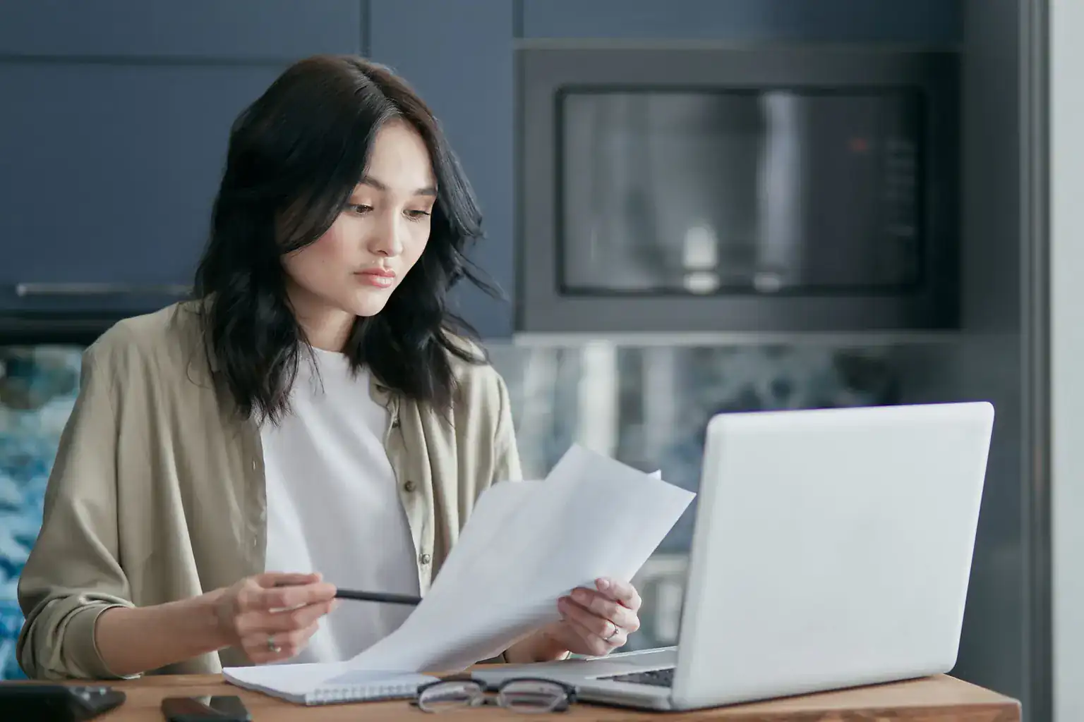 A woman at her kitchen table reading through paperwork with a pen beside an open laptop