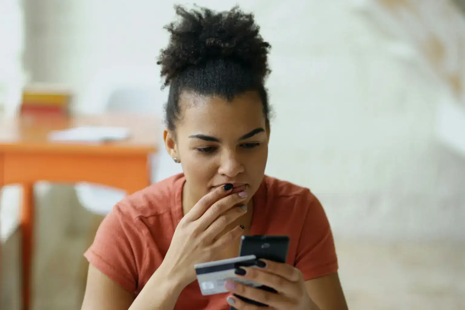 A young woman looks thoughtfully at her phone while holding a bank card at home