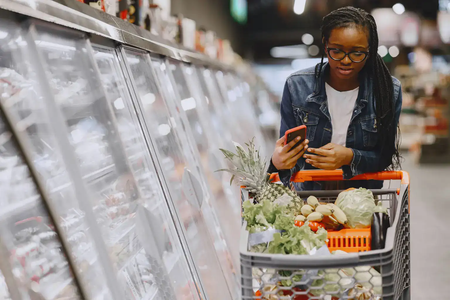 A woman checks her phone while pushing a trolley of fresh vegetables past supermarket chiller cabinets