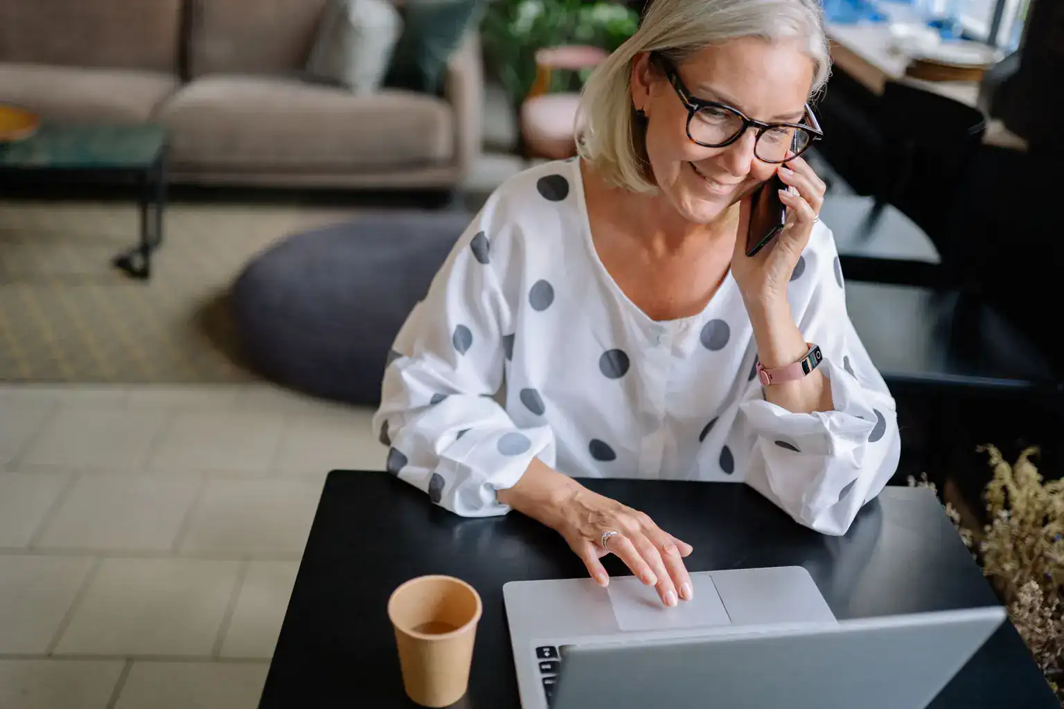 A smiling older woman talks on her phone while using a laptop at home