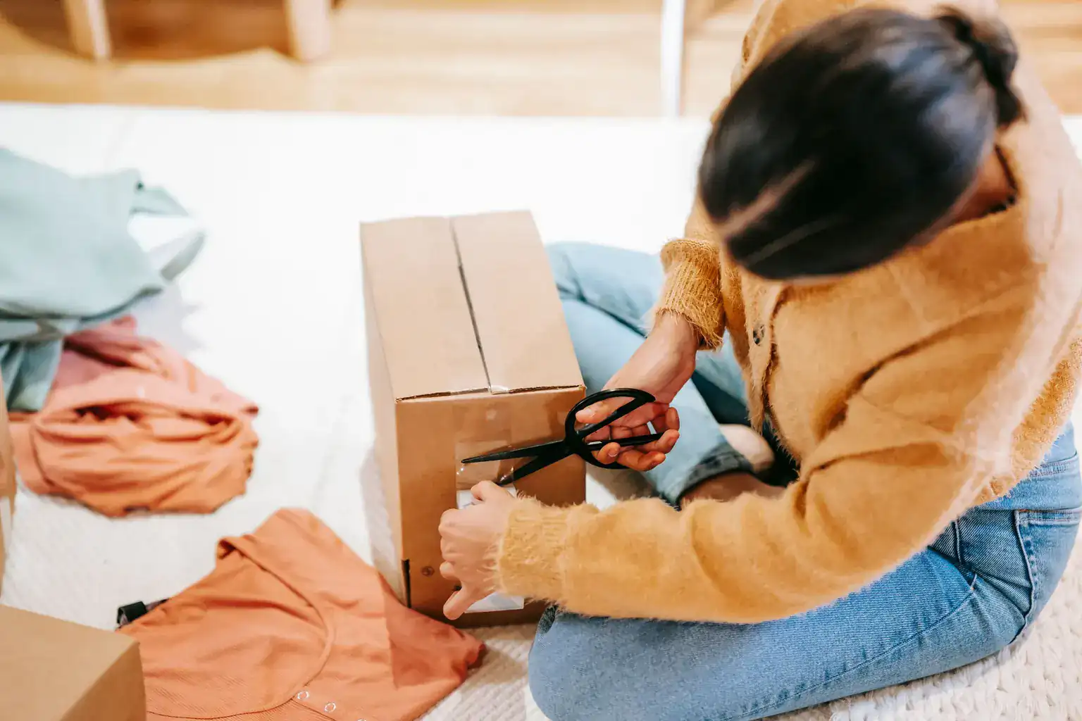 A woman sitting on the floor at home taping up a cardboard box of clothes to post