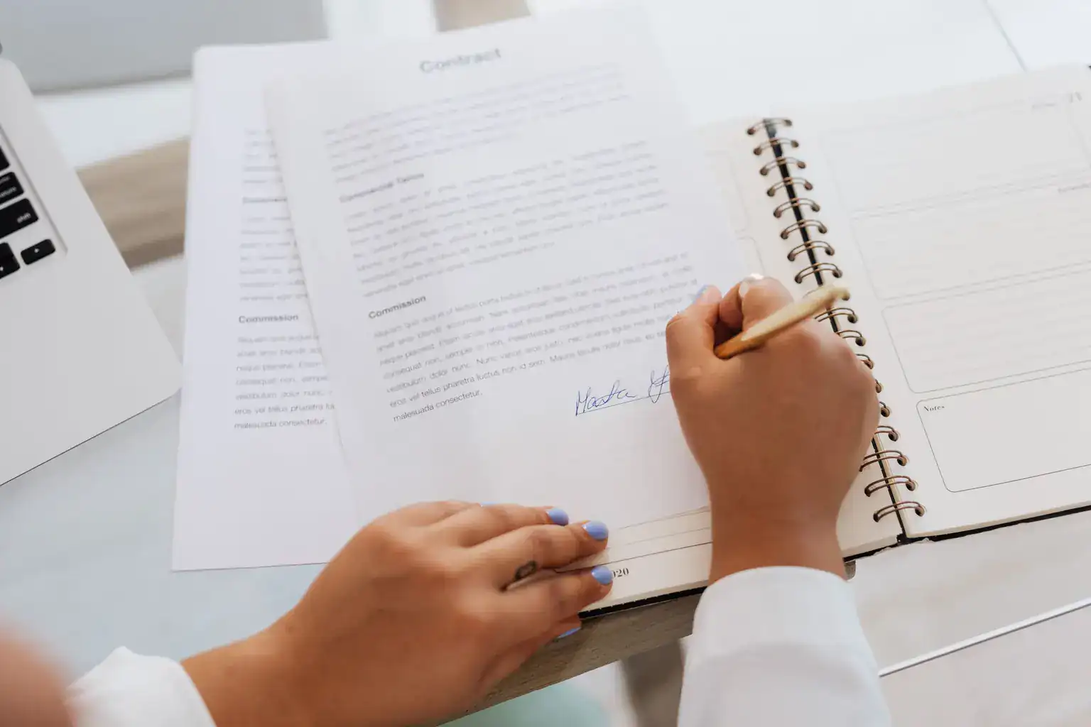 A woman signs the last page of a printed contract beside a notebook and laptop