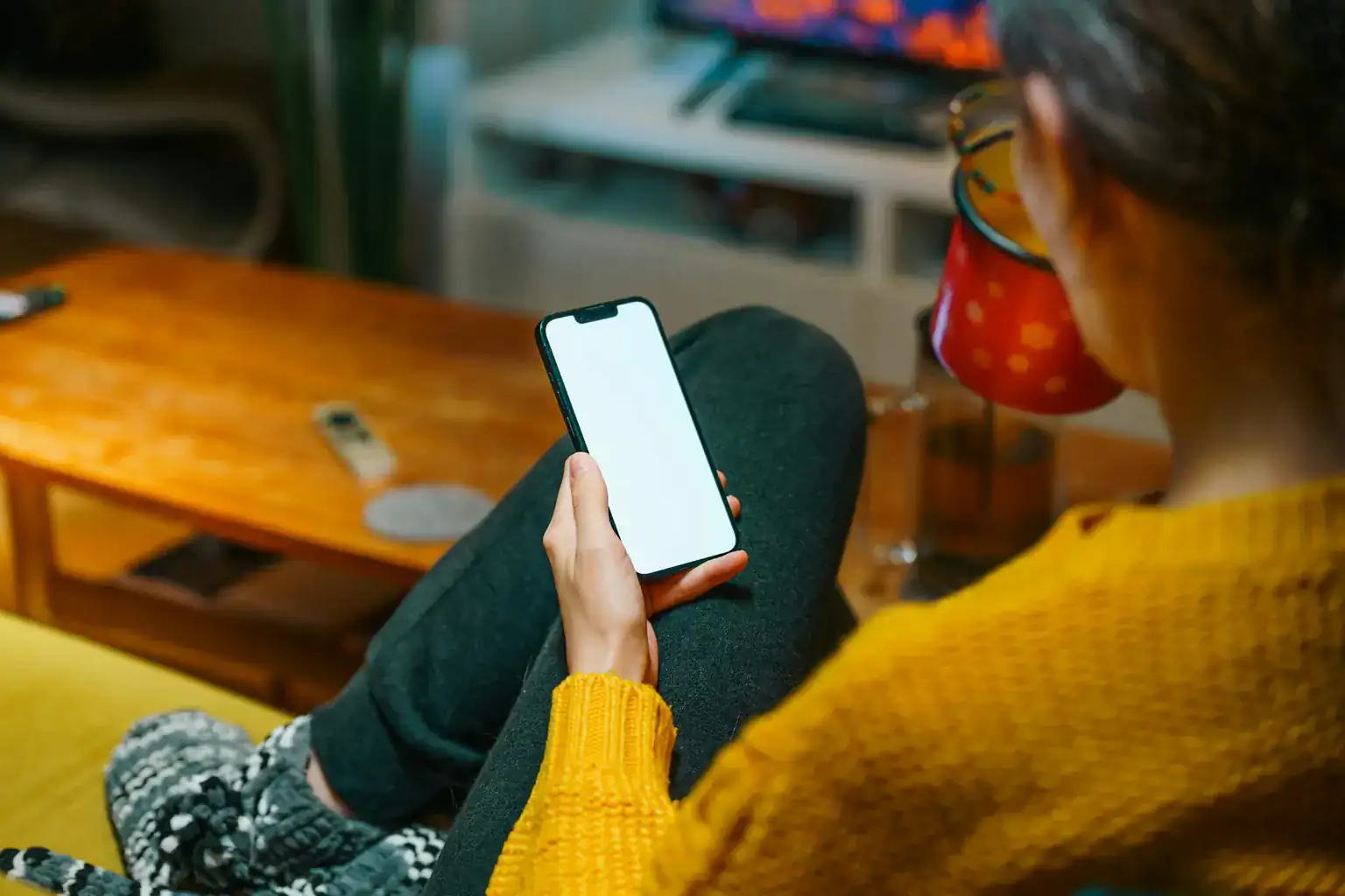 A woman in a mustard jumper relaxes on the sofa checking her phone with a mug of tea