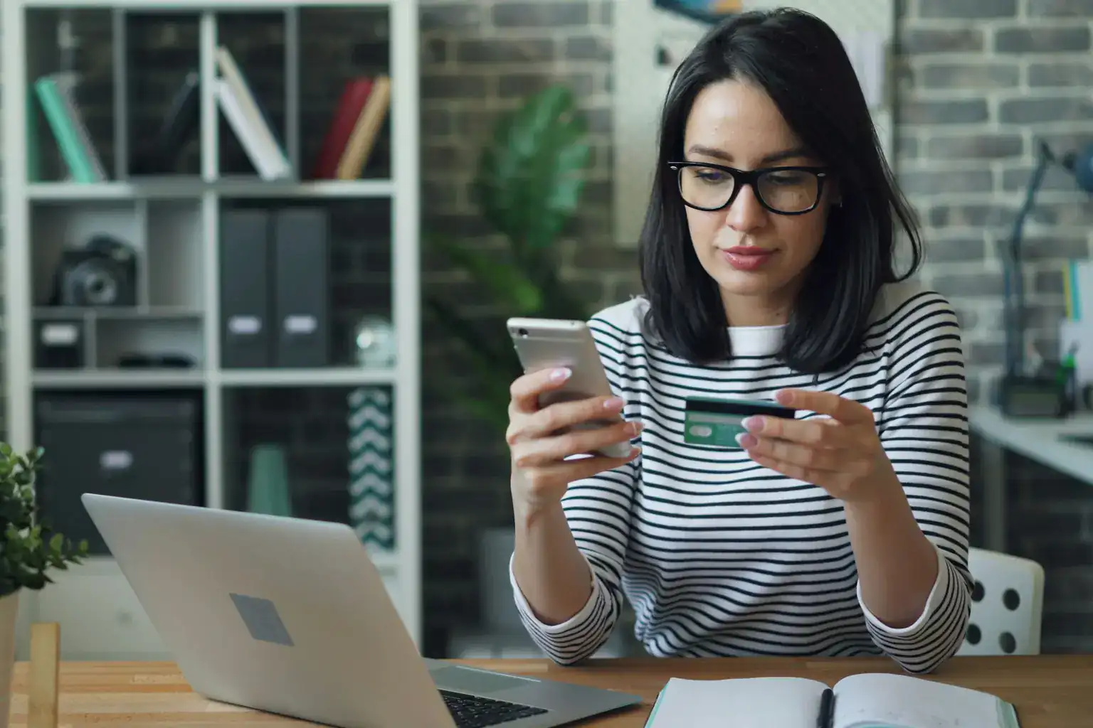 A woman at a home desk holds a bank card while checking her phone